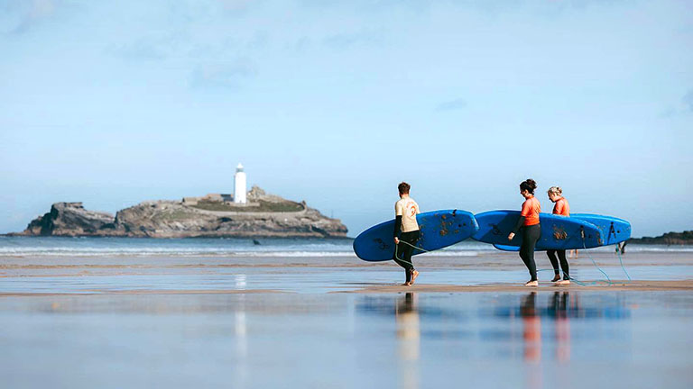 Student surfers walking across Gwithian beach with surfboards, and Godrevy Lighthouse in the background
