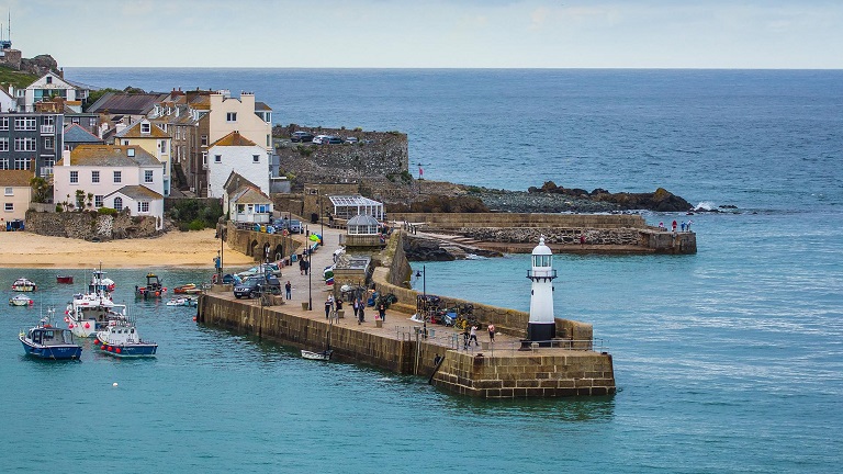 A scene showing St Ives harbour and lighthouse near high tide with fishermen's cottages in the background. 