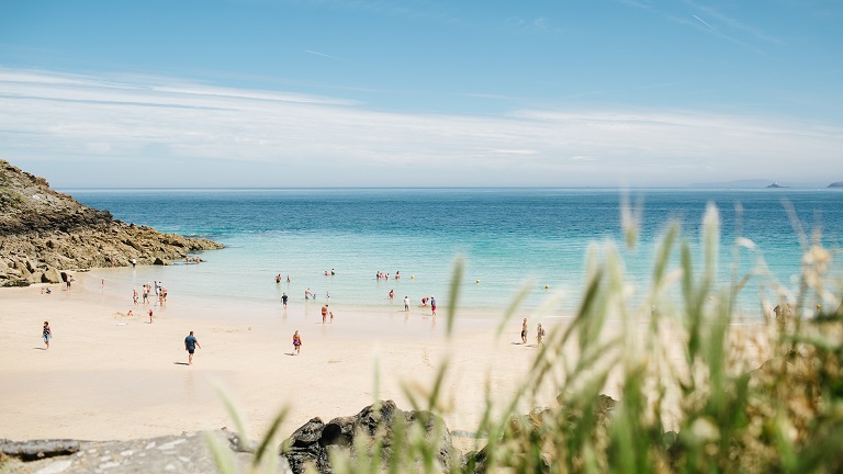 A stunning view of the white sand and turquoise sea of St Ives, with people enjoying their time on the beach in the background and marram grass in the foreground. 