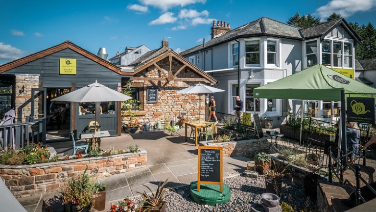 An outside view of Granny Dowbekin's riverside cafe with its tables and umbrellas outside in the sunshine, located in Pooley Bridge near Penrith in Cumbria