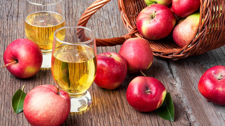 Two glasses of cider surrounded by apples tumbling out of a wicker basket. You can enjoy cider tasting at Sandford Orchard in Crediton, Devon. 