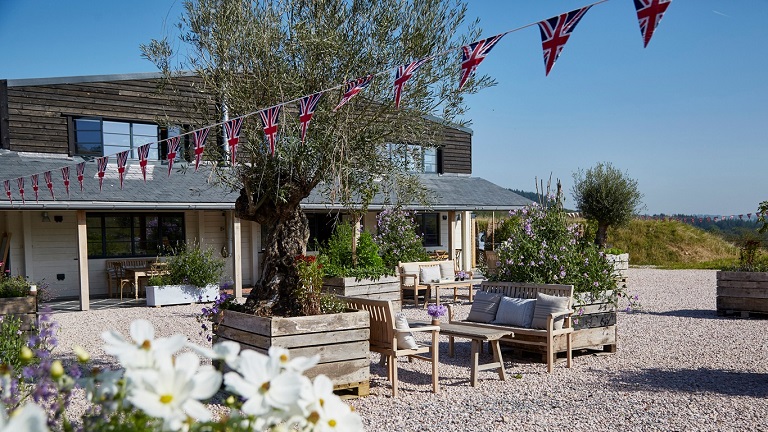 An exterior view of Heron Valley farm and orchard's shop, restaurant and bar in Loddiswell, Kingsbridge, decorated with flags and peppered with flowers and shrubs