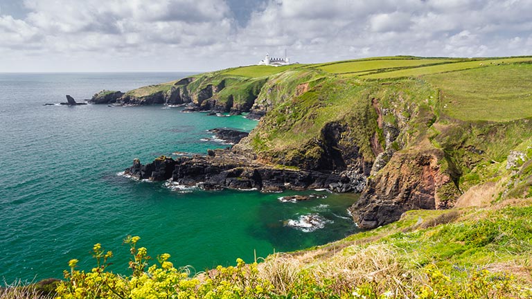 Rugged cliffs and green fields tower above the rocky cove at Housel Bay in Cornwall