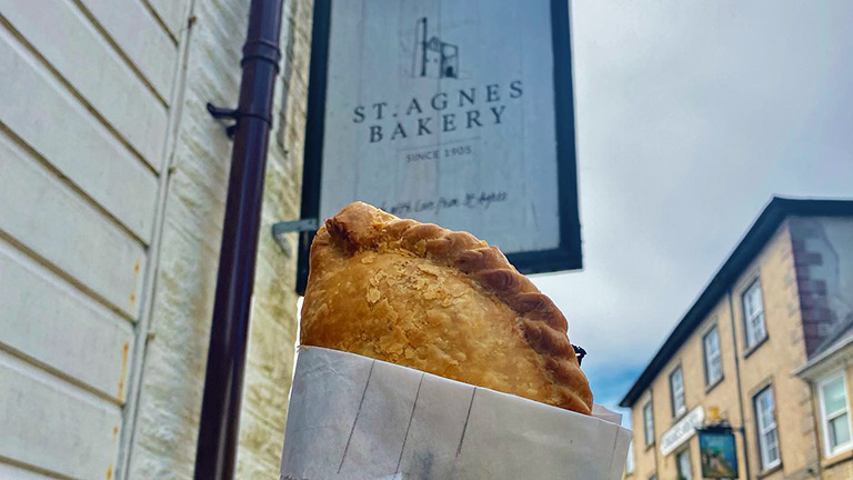 A golden crusted pasty held up underneath the sign for St Agnes Bakery