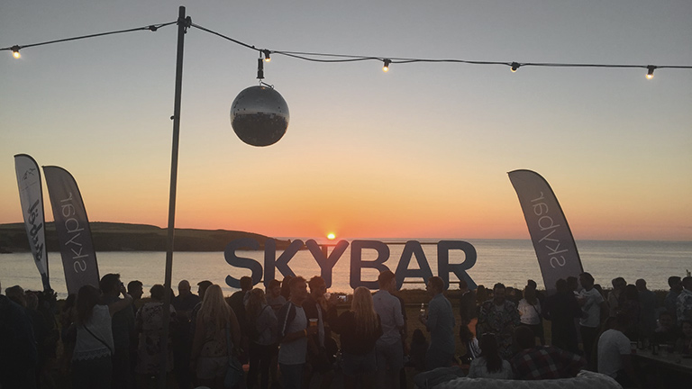 A sign of 'Sky Bar' illuminated by a setting sun over the sea on the Cornish coast