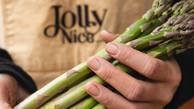A hand holding a fresh bunch of asparagus with a Jolly Nice apron behind