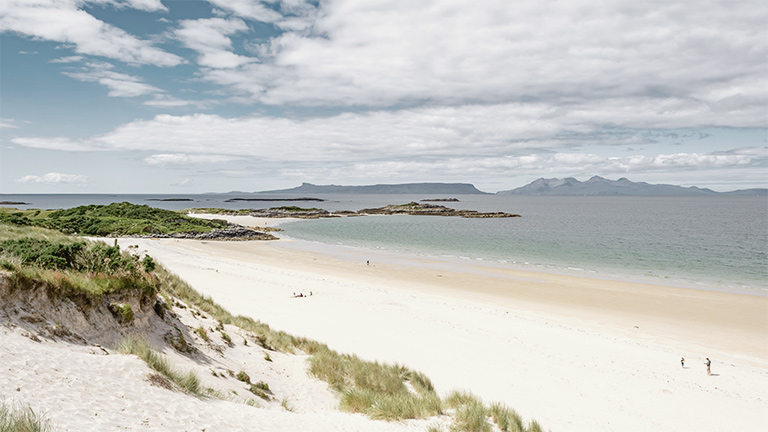 The white sand and soft dunes of Camusdarach Beach in Scotland with the sea and mountains beyond