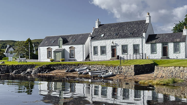 A view of Praban Na Linne Gaelic whiskies building reflected in mirror-like loch waters on a sunny day in Skye