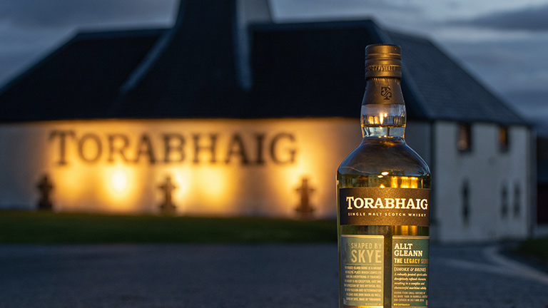 A bottle of Torabhaig Skye whisky with the distillery illuminated in the evening light behind