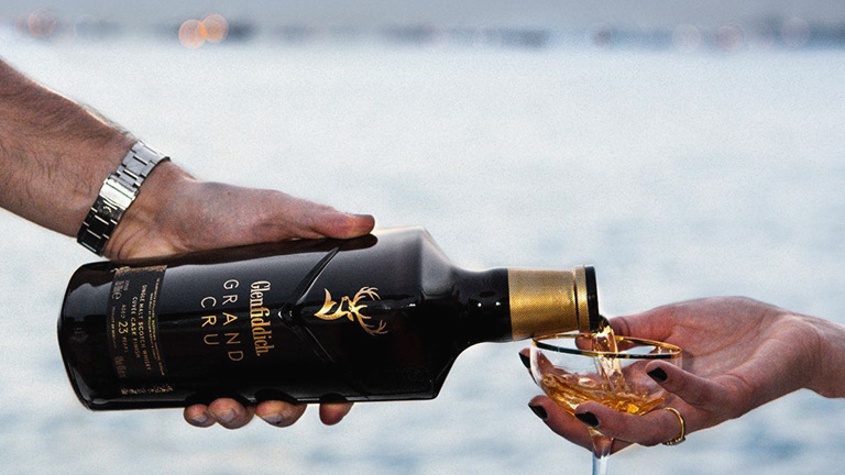 A glass of Glenfiddich Grand Cru being poured into a pretty glass with a lakeside backdrop