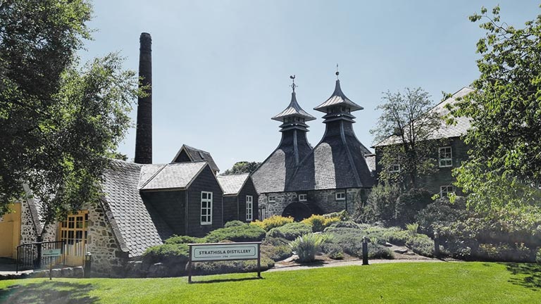 The sun-kissed exteriors of Strathisla Distillery framed by greenery and topped by blue skies