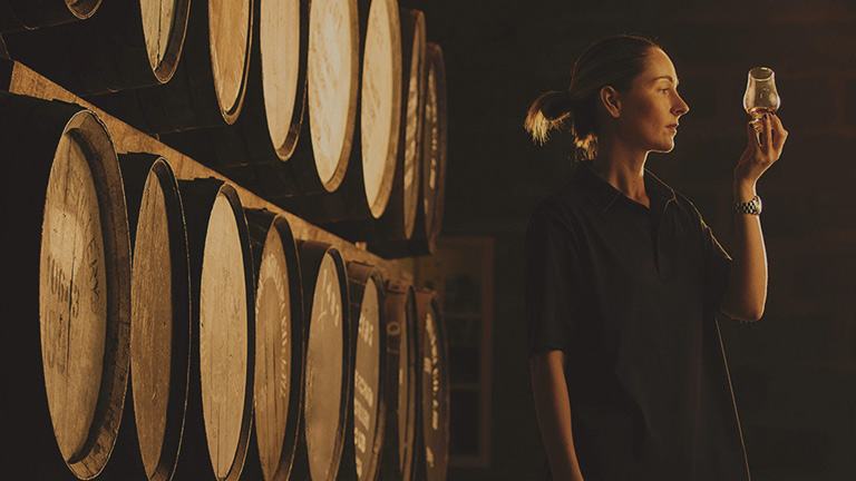 A whisky distiller inspecting a glass of whisky next to oak barrels at Glen Grant Distillery