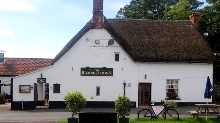 The white-washed frontage of the thatched Bicknoller Inn pub with a bicycle sign in front