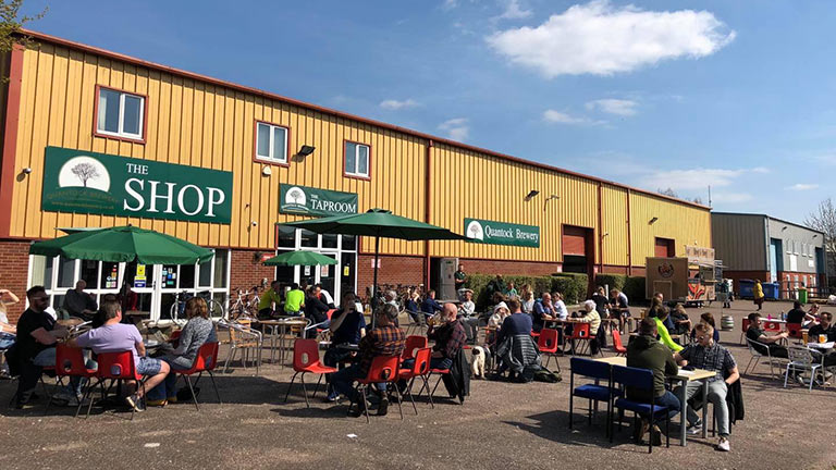 Groups of people sat around tables and chairs in the sunshine outside the Quantock Brewery in Bishops Lydeard